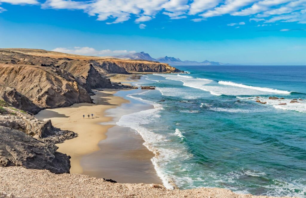 Surfers walk along a Canary Islands sand beach with rocky cliffs in background.